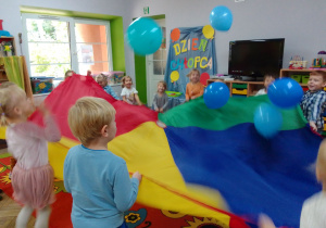 Dzieci poruszają chustą animacyjną, nad którą unoszą się niebieskie balony.