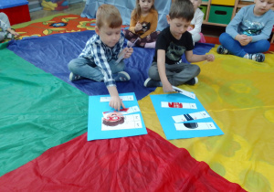 Antek i Mikołaj siedzą na kolorowej chuście i układają puzzle z bohaterami bajek. Wokół siedzą dzieci i dopingują kolegów.
