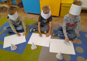 Trzech chłopców siedzi na dywanie i rysuje z zakrytymi oczami wymarzone auta na białych kartkach. Przed kartkami leżą czapeczki.