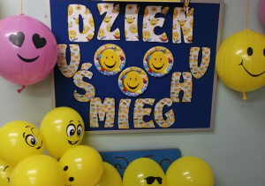 Na granatowej tablicy umieszczony jest napis Dzień Uśmiechu oraz uśmiechnięte żółte buźki. Obok tablicy wiszą balony w kolorze żółtym i różowym z uśmiechniętymi buziami. Na białym stoliku leżą czapeczki, dyplomy, odznaki oraz żółte balony z uśmiechniętymi buziami.