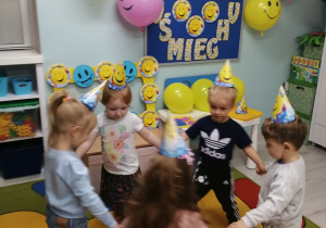 Zabawa taneczna w kole. Dzieci na głowach mają założone papierowe czapeczki z uśmiechniętą buzią. W tle dekoracja z okazji Dnia Uśmiechu.
