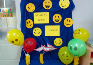 Dekoracja na tablicy: napis – Światowy Dzień Uśmiechu, na niebieskim tle żółte uśmiechnięte buzie z papieru oraz "uśmiechnięte” kolorowe balony.