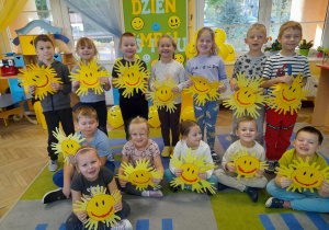 Dzieci z radością prezentują wykonane przez siebie słoneczka. W tle tablica z napisem „Dzień Uśmiechu”, okno oraz kącik domowy.