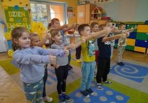 Dzieci bawią się przy piosence „Chu chu ua”. Stoją na dywanie z wyciągniętymi do przodu rękami i kciukami podniesionymi do góry.