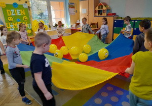 Dzieci idą w kole na dywanie i trzymają chustę animacyjną, na której leżą żółte balony z uśmiechniętymi buźkami.