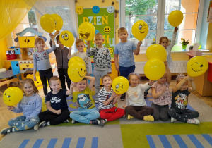 Dzieci z radością pozują do zdjęcia z żółtymi balonami. Na kilku balonach widać uśmiechnięte buźki.