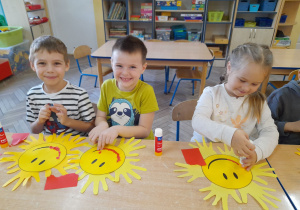 Hubert, Krzyś i Alicja siedzą przy stole obok siebie, a przed nimi leżą uśmiechnięte słoneczka. Dzieci wydzierają z czerwonego papieru i wyklejają uśmiech.