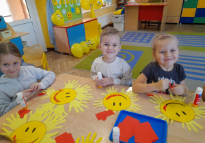 Amelka, Oskar i Gabrysia uśmiechają się podczas wykonywania pracy plastycznej przy stoliku. Dzieci siedzą obok siebie i wyklejają wydzieranką z czerwonego papieru uśmiech słoneczka.