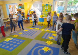 Na dywanie leżą w półkolu wykonane przez dzieci słoneczka. Przedszkolaki w parach tańczą przy piosence „Wesołe słoneczko”. W tle dekoracja z okazji Dnia Uśmiechu, okna, kącik domowy, przyrodniczy.