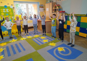 Dzieci stoją na dywanie, a przed nimi leżą wykonane słoneczka z kartonu. Przedszkolaki podnoszą do góry ręce naśladując promyczki słońca przy piosence „Wesołe słoneczko”.