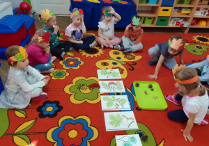 Dzieci w opaskach na głowach siedzą na dywanie w półkolu, przed nimi ilustracje liści i owoców z różnych drzew.