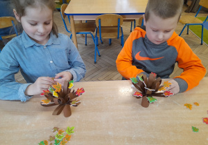 Alicja i Bruno siedzą przy stole i wykonują drzewko jako element dekoracji. Dzieci naklejają jesienne listki.
