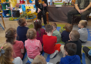 Dzieci siedzą na dywanie i z uwagą słuchają Pani edukator, która opowiada dzieciom o dębie pokazując gałązkę z brązowymi liśćmi. Obok siedzi Pan podleśniczy.