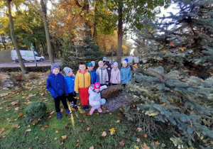 Dzieci pozują do zdjęcia przy rzeźbie szyszki na tle jesiennych drzew.