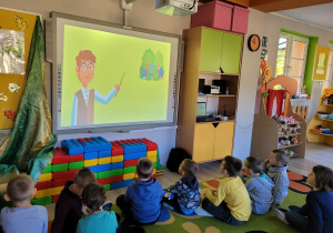 Dzieci z grupy siedzą na dywanie przed tablicą multimedialną i oglądają film edukacyjny