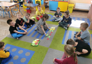 Dzieci siedzą w kole na dywanie. W środku koła Ala K. układa ciąg rytmów z wykorzystaniem szablonów figur geometrycznych.