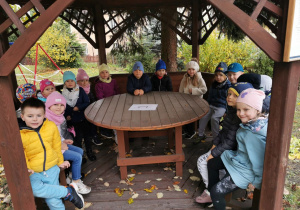 Dzieci z grupy "Słoneczek" siedzą w altanie ogrodowej. Na stole widać kartkę z cyfrą 1. Przedszkolaki słuchają objaśnień do wykonania pierwszego zadania.