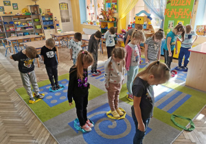 Dzieci z grupy "Słoneczka" stoją na dywanie na którym rozłożone są szarfy. Dzieci zajmują miejsce w szarfach.