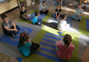 Dzieci siedzą na dywanie po turecku w trakcie zabawy z woreczkami - woreczki leżą za nimi.