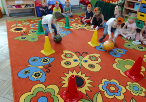 Antek i Franek z opaskami na głowach turlają dynie między pachołkami, reszta grupy siedzi na dywanie i kibicuje kolegom, z tyłu kąciki zabaw.