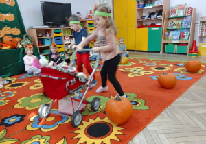 Mikołaj i Lena prowadzą dziecięce wózki z lalkami slalomem między dyniami rozstawionymi na dywanie. Z tyłu: dekoracja, telewizor i kąciki zabaw.
