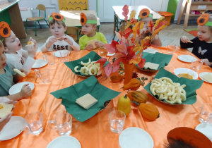 Motylki w opaskach na głowach siedzą przy stole, na którym są: chrupki, wafelki, mus dyniowo-jabłkowy, szklanki z wodą, ozdobne dynie i kolorowe jesienne liście.