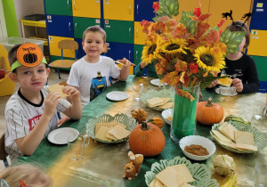 Dzieci z grupy siedzą przy stole w trakcie dyniowego poczęstunku. Na stole widać wafle, konfiturę dyniową, dżem jabłkowo – dyniowy, wazon z jesiennymi kwiatami i małe ozdobne dynie.