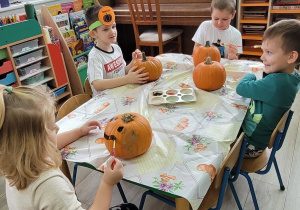 Dzieci z grupy siedzą przy stoliku i malują dynie farbami.