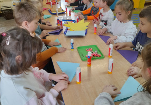 Dzieci siedzą przy połączonych stolikach i składają kolorowe kartony. Na stolikach leżą materiały potrzebne do wykonania pieska techniką origami. W tle tablica z napisem "Dzień Kundelka", kącik domowy, przyrodniczy.