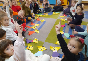 Dzieci siedzą na dywanie w kole, a przed nimi leżą pieski wykonane techniką origami. Przedszkolaki wymyśliły imię dla swojego pupila i podnoszę rękę do góry chcąc udzielić odpowiedzi.