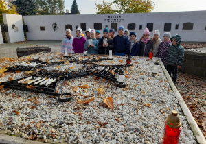 Dzieci stoją obok Pomnika Bohaterów Walk nad Bzurą. Alicja i Wiktoria trzymają w rękach znicze. W tle "Mur pamięci" z tablicami pamiątkowymi.
