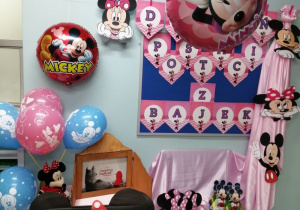Granatowa tablica, na której znajduje się napis "Dzień Postaci z Bajek". Obok tablicy na różowym materiale przypięte są postacie Myszki Mickey. Na szafce okrytej różowym materiałem leżą papierowe czapeczki, odznaki oraz niebieskie i różowe bajkowe balony. Obok szafki stoi Teatrzyk Kamishibai oraz "fotobudka Myszki Mickey".