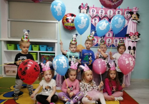 Zdjęcie grupowe na tle dekoracji z okazji Dnia Postaci z Bajek. Dzieci trzymają balony w kolorze niebieskim i różowym z Myszką Mickey.
