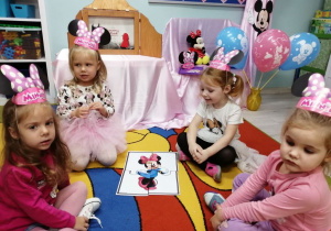 Grupka dziewczynek siedzi na dywanie, a przed nimi leży ułożony z części obrazek z Myszką Mickey.