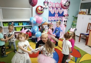 Zabawa taneczna przy muzyce w kółeczkach. Dzieci na głowach mają założone papierowe czapeczki Myszki Mickey. W tle dekoracja z okazji Dnia Postaci z Bajek.