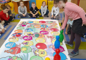 Dzieci siedzą na dywanie wokół gry planszowej "Bal u Osiołka". Alicja po wyrzuceniu kostką przesuwa zielony kręgiel po kolorowych balonikach z bajkowymi postaciami.