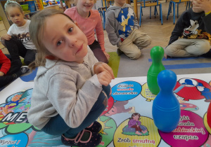 Amelka przykucnęła na planszy z grą "Bal u Osiołka" i próbuje zrobić smutną minę zgodnie z poleceniem napisanym na baloniku. Obok dziewczynki stoją dwa kręgle, a z tyłu siedzi kilkoro dzieci.