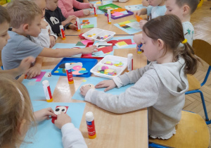 Dzieci siedzą przy połączonych stołach i wykonują bajkowe postacie - naklejają na niebieskie kartki elementy w kształcie figur geometrycznych.