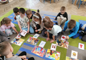 Dzieci siedzą na dywanie, a przed nimi leżą bajkowe obrazki. Przedszkolaki dopasowują rekwizyty do odpowiednich bajek. Krzyś trzyma obrazek z wrzecionem.