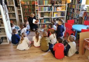 Dzieci siedzą razem z panią z biblioteki, która prezentuje różne książki z bajkami.
