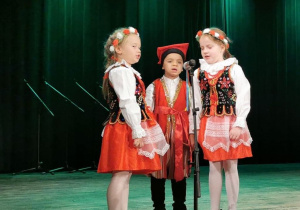Alicja, Tymon i Alicja przebrani w stroje krakowskie stoją na scenie przed mikrofonem i śpiewają piosenkę "Jestem Polakiem".