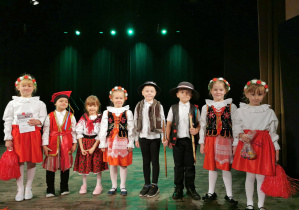 Ośmioro dzieci stoi na scenie obok siebie i pozuje do zdjęcia po występie. Alicja trzyma dyplom dla zespołu "Stokrotka", a Oliwia słodycze.