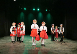 Przedszkolaki stoją na scenie. Na pierwszym planie Alicja i Oliwia trzymają pompony w kolorze czerwonym i mikrofony w rękach. Dziewczynki ubrane są w białe bluzki i czerwone spódnice, a na głowach mają biało-czerwone wianki. Z prawej strony stoją Gabryś, Zosia i Krzyś w strojach góralskich, a z lewej strony Alicja, Tymon i Alicja w strojach krakowskich.
