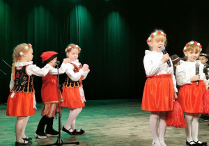 Alicja i Oliwia stoją obok siebie na scenie. Alicja śpiewa piosenkę do mikrofonu, a z lewej strony troje dzieci przebranych w stroje krakowskie jedzie w pociągu naśladując treść piosenki.