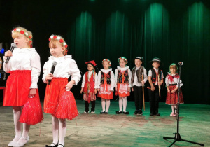 Alicja śpiewa piosenkę do mikrofonu, a obok stoi Oliwia. Dziewczynki ubrane są w białe bluzki i czerwone spódnice, a na głowach mają biało-czerwone wianki. W tle sześcioro dzieci przebranych w stroje krakowskie i góralskie stoi obok siebie.