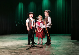 Krzyś, Zosia i Gabryś ubrani w stroje góralskie stoją na scenie przed mikrofonem i śpiewają piosenkę "Jestem Polakiem"..