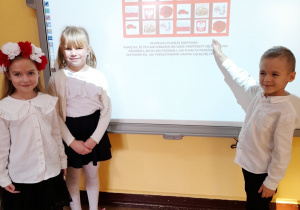Vanessa, Alicja i Oskar stoją przed tablicą multimedialną i prezentują łączące się z Polską sudoku obrazkowe.