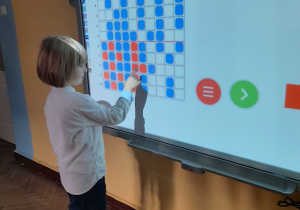 Maksymilian stoi przed wyświetloną na ekranie tablicy multimedialnej kratownicą i zaznacza na kolor czerwony pola na podstawie podanych koordynatów.