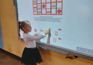Gabrysia układa sudoku obrazkowe na tablicy multimedialnej. Dziewczynka przeciąga obrazek w określone miejsce zgodnie z podanym warunkiem.