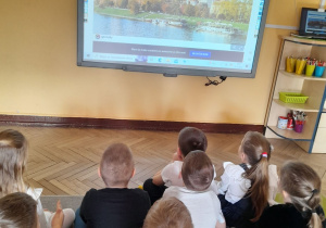 Dzieci siedzą na dywanie przed tablica multimedialna, na której wyświetlony jest temat "Polska moja ojczyzna".